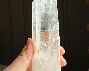 May include: A clear quartz crystal point, held in a hand. The crystal is transparent with visible internal structures and a sharp, pointed tip. The quartz has a rough, natural texture and a few golden inclusions.