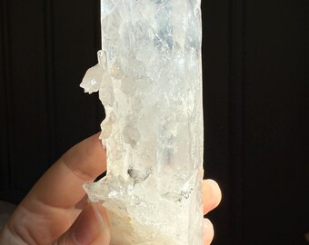 May include: A clear quartz crystal point with a faceted top and a rough, textured surface. The crystal is held in a hand, with sunlight illuminating its translucent structure. The crystal is a natural, raw mineral specimen.