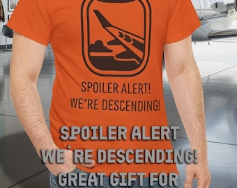 May include: An orange t-shirt with a black graphic of an airplane window and the text "SPOILER ALERT! WE'RE DESCENDING! GREAT GIFT FOR AVIATION LOVERS." A man is wearing the shirt in front of a hangar with a jet.