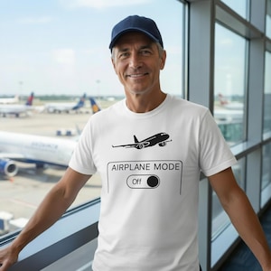 May include: White t-shirt with a black airplane graphic and the text "AIRPLANE MODE Off". The shirt is worn by a person in front of a window with airplanes in the background. The person is wearing a navy blue baseball cap.