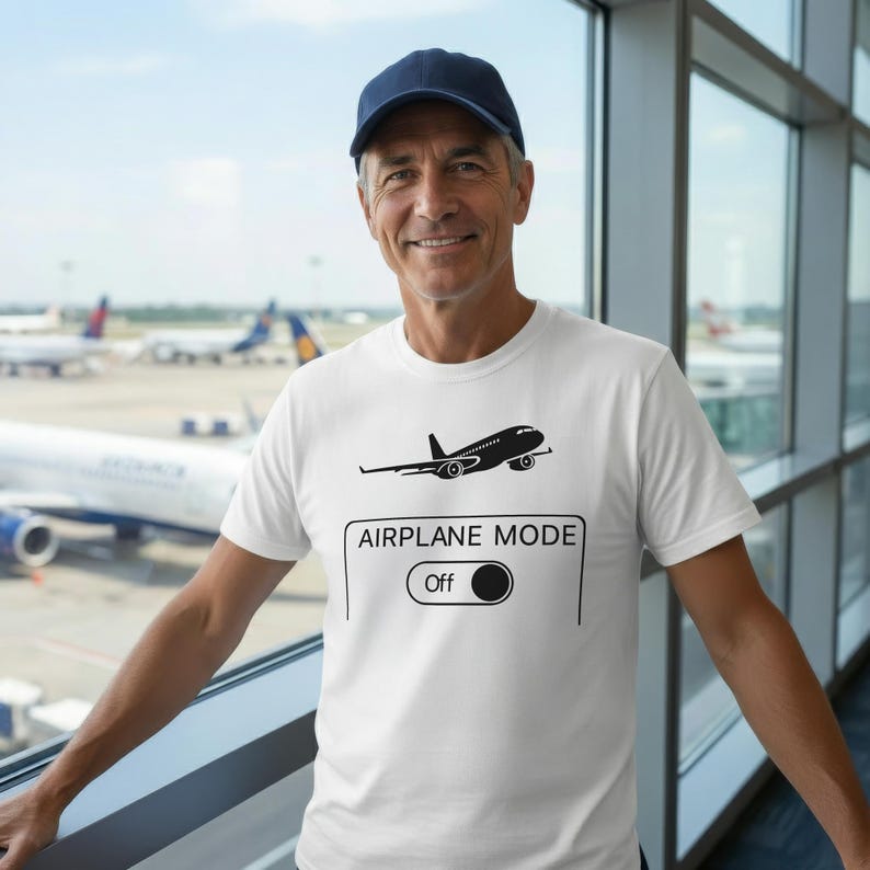 May include: White t-shirt with a black airplane graphic and the text "AIRPLANE MODE Off". The shirt is worn by a person in front of a window with airplanes in the background. The person is wearing a navy blue baseball cap.