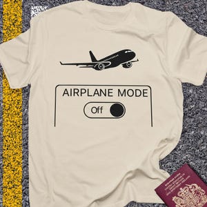 May include: A cream-colored t-shirt with a black graphic of an airplane and the words "AIRPLANE MODE" above an "Off" switch. The shirt is slightly wrinkled, suggesting it is made of a soft material.