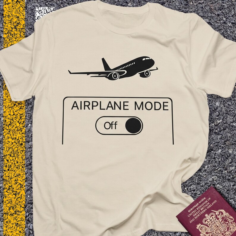 May include: A cream-colored t-shirt with a black graphic of an airplane and the words "AIRPLANE MODE" above an "Off" switch. The shirt is slightly wrinkled, suggesting it is made of a soft material.