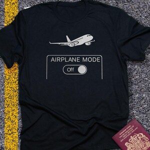 May include: Black t-shirt with a white graphic of an airplane and the words "AIRPLANE MODE Off". A passport is visible in the bottom right corner. The shirt is on a textured surface.