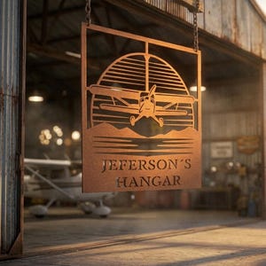 May include: A rectangular metal sign with a rust-colored finish, featuring a cutout design of a small airplane flying in front of a sun with horizontal lines. Below the airplane, the text JEFERSON'S HANGAR is cut out from the metal surface.