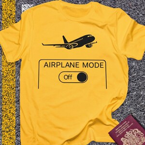 May include: Yellow t-shirt with a black airplane graphic and the words "AIRPLANE MODE Off". The design includes a black airplane silhouette above a rectangular box with a toggle switch. A passport is visible in the bottom right corner.