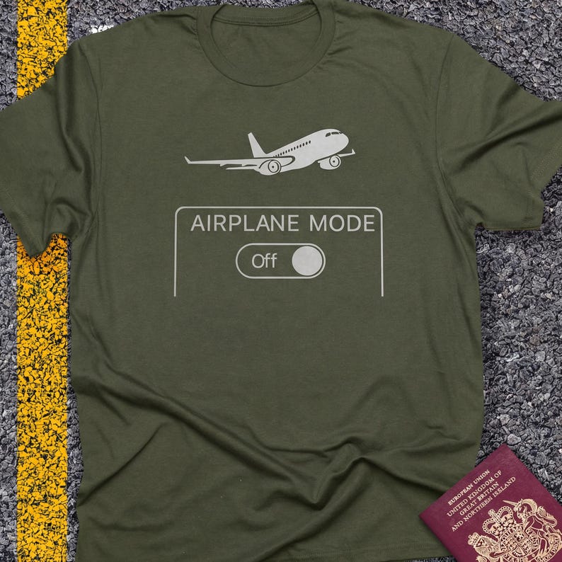 May include: Olive green t-shirt with a white graphic of an airplane and the words "AIRPLANE MODE" above a switch labeled "Off". The shirt is made of a soft material.