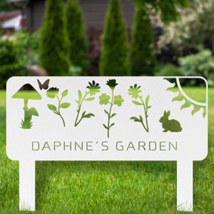 Puede incluir: Letrero de jard&iacute;n de metal blanco con dise&ntilde;os recortados en verde de una seta, mariposa, flores, sol y conejo. El texto "DAPHNE'S GARDEN" se muestra en verde. El letrero est&aacute; en un jard&iacute;n con c&eacute;sped.