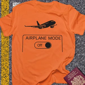 May include: An orange t-shirt with a black graphic of an airplane and the words "AIRPLANE MODE" above an "Off" switch. The shirt is made of a soft material and is suitable for casual wear.