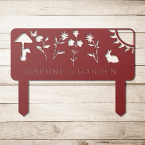 Puede incluir: Un letrero de jard&iacute;n de metal rojo con dise&ntilde;os recortados de una seta, flores, una mariposa, un conejo y un sol. El texto "DAPHNE'S GARDEN" est&aacute; grabado. El letrero tiene dos estacas para colocarlo en el suelo.