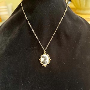 May include: A gold-colored chain necklace with a cameo pendant. The pendant features a black and white profile of a person in an oval frame with a decorative border. The necklace is displayed on a black surface.