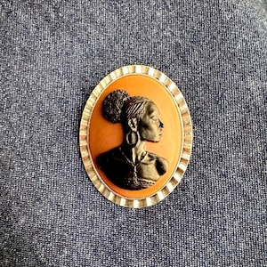 May include: Oval-shaped cameo brooch featuring a profile of a woman with her hair up in a bun and wearing hoop earrings. The cameo is set against an orange background and framed by a silver-colored, ridged border. The brooch is a vintage style.