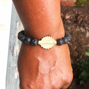 May include: A black lava rock bracelet with a gold charm. The charm has a detailed design and is in the center of the bracelet.