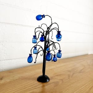 May include: A black metal wire tree sculpture featuring multiple small, cobalt blue glass bottles hanging from its branches. The tree has a solid circular base and thin, twisted wire limbs that support the various shapes of blue glass vessels.
