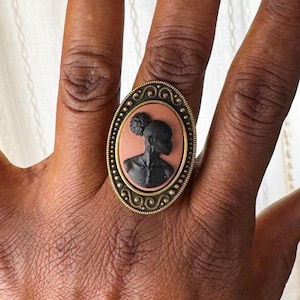 May include: Oval-shaped ring with a cameo design. The ring features a black silhouette of a person with a bun hairstyle against a peach background. The ring has a decorative gold-colored frame with a beaded edge.