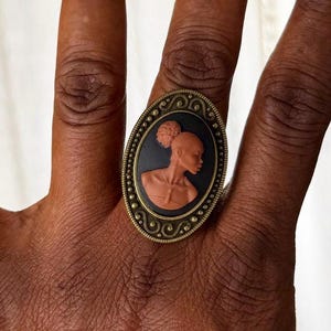 May include: Oval-shaped ring with a cameo design. The ring features a black background with a profile of a woman in a peach color. The ring has a decorative, antique-style brass frame.
