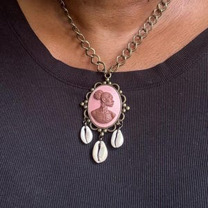 May include: A bronze-toned necklace featuring a pink oval pendant with a woman's profile in relief. The pendant is adorned with a decorative frame and three dangling white seashells. The necklace is worn against a dark top.