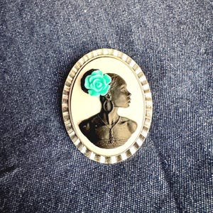 May include: Oval-shaped cameo brooch featuring a profile of a woman with a turquoise flower in her hair. The cameo is set in a silver-toned frame with a striped border. The woman is wearing hoop earrings and a necklace.