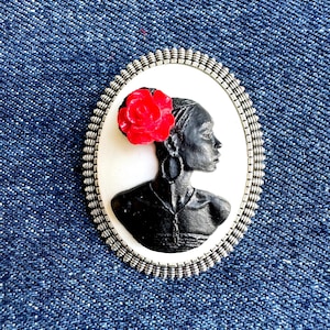 May include: Oval-shaped brooch featuring a black profile of a person with a red rose in their hair. The profile is set against a white background, framed by a silver-colored, textured border. The brooch is set on a blue denim background.