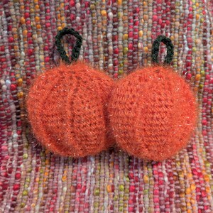Può includere: Due ornamenti a forma di zucca arancione lavorati a maglia con steli verdi e accenti glitterati. Le zucche sono rotonde e soffici, su uno sfondo colorato e tessuto. Questi oggetti decorativi sono perfetti per l'autunno o Halloween.