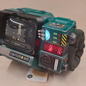 May include: A teal and black wearable wrist-mounted computer replica featuring a screen displaying a character, a glowing blue light, a red illuminated mesh panel, various knobs, and a label reading 27OCT2102 0634 with a bell icon.