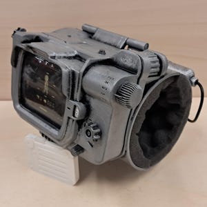 May include: A grey, weathered wrist-mounted computer replica featuring a rectangular screen with visible interface graphics, multiple side-mounted adjustment knobs, and a black foam-lined circular opening for the arm, finished with metallic paint.