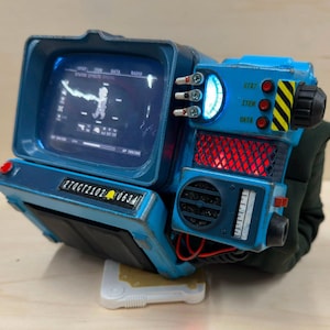 Op de afbeelding: Een blauwe polscomputer met een scherm dat een personagepictogram toont, drie tuimelschakelaars, een oplichtende blauwe meter, rode indicatielampjes en een zwarte draaiknop. Het apparaat heeft een label met 27OCT2102 en gele waarschuwingsstrepen.