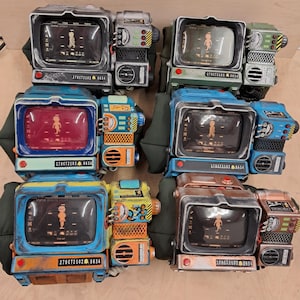 May include: Six wearable wrist-mounted computer replicas featuring rectangular screens displaying a character icon, various dials, buttons, and mesh vents. Each unit is finished in different colors including metallic silver, green, blue, and copper tones.