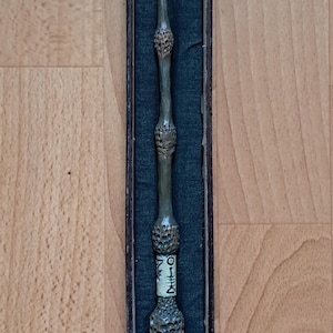 Peut inclure: Une baguette détaillée, probablement un accessoire ou un objet de collection, est présentée dans un coffret. La baguette est de couleur vert-brun sourd avec des détails complexes le long de sa longueur, y compris des sections bulbeuses et une poignée texturée. La boîte est sombre avec un intérieur turquoise.