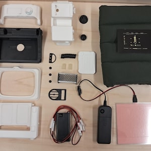 May include: A collection of disassembled electronic components including a dark green quilted fabric cover, white and black plastic housing parts, a small metal mesh screen, a black battery pack with red and black wires, and a small circuit board.