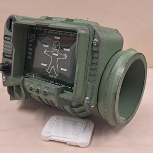 Könnte beinhalten: Eine salbeigrüne Pip-Boy-Nachbildung mit einem digitalen Bildschirm, der einen Vault-Boy-Charakter, Statistiken sowie den Text "STATS", "SPECIAL", "Skills" und "Perks" anzeigt. Ein kleines weißes rechteckiges Zubehörteil liegt vor dem Gerät.