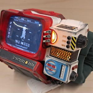 May include: A red and white wearable wrist computer featuring a digital screen displaying a character icon and the text CountryRoads147. The device includes a glowing orange gauge, black knobs, a blue circular heart icon, and a metal plate labeled 27OCT2102 0634.