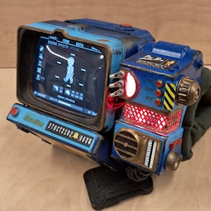 May include: A blue wrist-mounted computer prop featuring a screen displaying a character icon and the text 'pigeon_Hooties'. The device includes red illuminated panels, a 'Nuka-Cola Quantum' label, and a '270CT2102 0634' serial number plate.