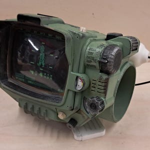 May include: A sage green wearable wrist-mounted computer replica featuring a dark screen with a glowing green character graphic, multiple textured control dials, metallic weathering details, and a black cable extending from the side of the device.