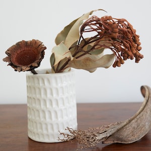 Puede incluir: Un jarrón de cerámica blanca con una superficie texturizada, que contiene flores y hojas secas marrones. El jarrón está sobre una superficie de madera.