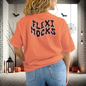 May include: A bright orange t-shirt with the text "Flexi Mocks" in black dripping letters. The shirt is being worn by a person with long brown hair and blue jeans.