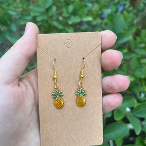 May include: Gold-tone pineapple earrings with yellow and green enamel detailing. The earrings are displayed on a brown card, with a green leafy background. The earrings have a hook closure.