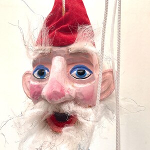 May include: A hand-carved wooden puppet of a gnome with a red pointed hat, blue eyes, and a long white beard. The gnome has a pink nose and mouth, and is wearing a blue shirt. White strings are attached to the puppet for manipulation.