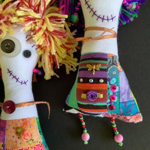 May include: Two fabric dolls with white bodies, yarn hair in yellow and purple, and button eyes. The dolls feature stitched mouths, colorful patchwork dresses with ribbon accents, metallic dragonfly charms, and dangling beaded legs.