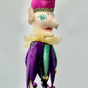 May include: A whimsical jester doll with a pink, textured hat and a purple and green patterned costume. The doll has a fabric face with painted features, a gold collar, and dangling bells. The jester is a decorative item.