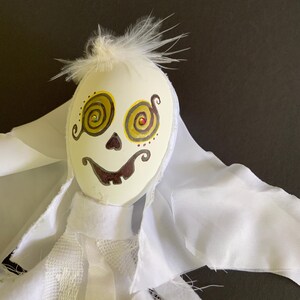 May include: A handmade Halloween ghost decoration with a painted face and white fabric. The ghost's face is oval-shaped, with yellow spiral eyes, a brown mouth, and a small tuft of white feathers on top. The ghost is wearing a white fabric shroud.