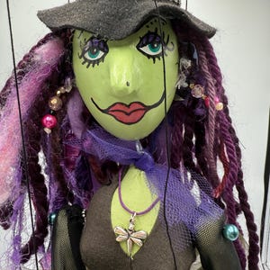 May include: A handmade witch puppet with a green face, red lips, and teal eyes. The puppet has purple and pink dreadlocks, a gray hat, and a dragonfly pendant. The witch is wearing a brown top and black mesh sleeves.