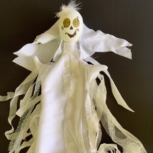 May include: A handmade white ghost decoration with a painted face featuring spiral eyes and a smiling mouth. The ghost has flowing fabric arms and a feathery top, perfect for Halloween or spooky decor.