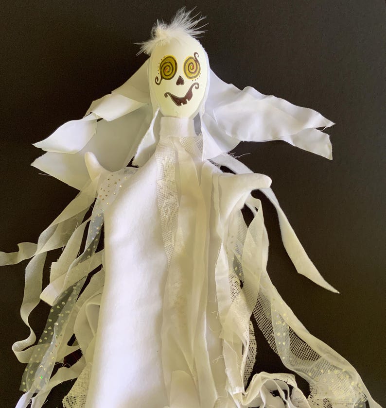 May include: A handmade white ghost decoration with a painted face featuring spiral eyes and a smiling mouth. The ghost has flowing fabric arms and a feathery top, perfect for Halloween or spooky decor.