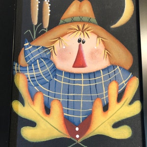 May include: Framed folk art painting of a scarecrow with a straw hat, blue plaid shirt, and a red nose. The background is black with a yellow crescent moon and cattails. The scarecrow has yellow leaves and a red and brown vest.