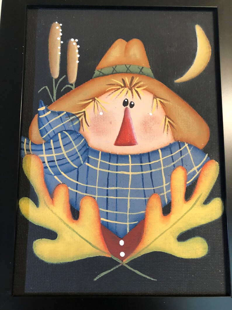 May include: Framed folk art painting of a scarecrow with a straw hat, blue plaid shirt, and a red nose. The background is black with a yellow crescent moon and cattails. The scarecrow has yellow leaves and a red and brown vest.