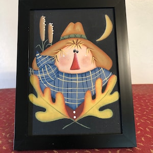 May include: Framed folk art painting of a scarecrow with a straw hat and blue plaid shirt. The scarecrow has a red nose and is surrounded by yellow leaves, cattails, and a crescent moon against a black background. The artwork is in a black frame.