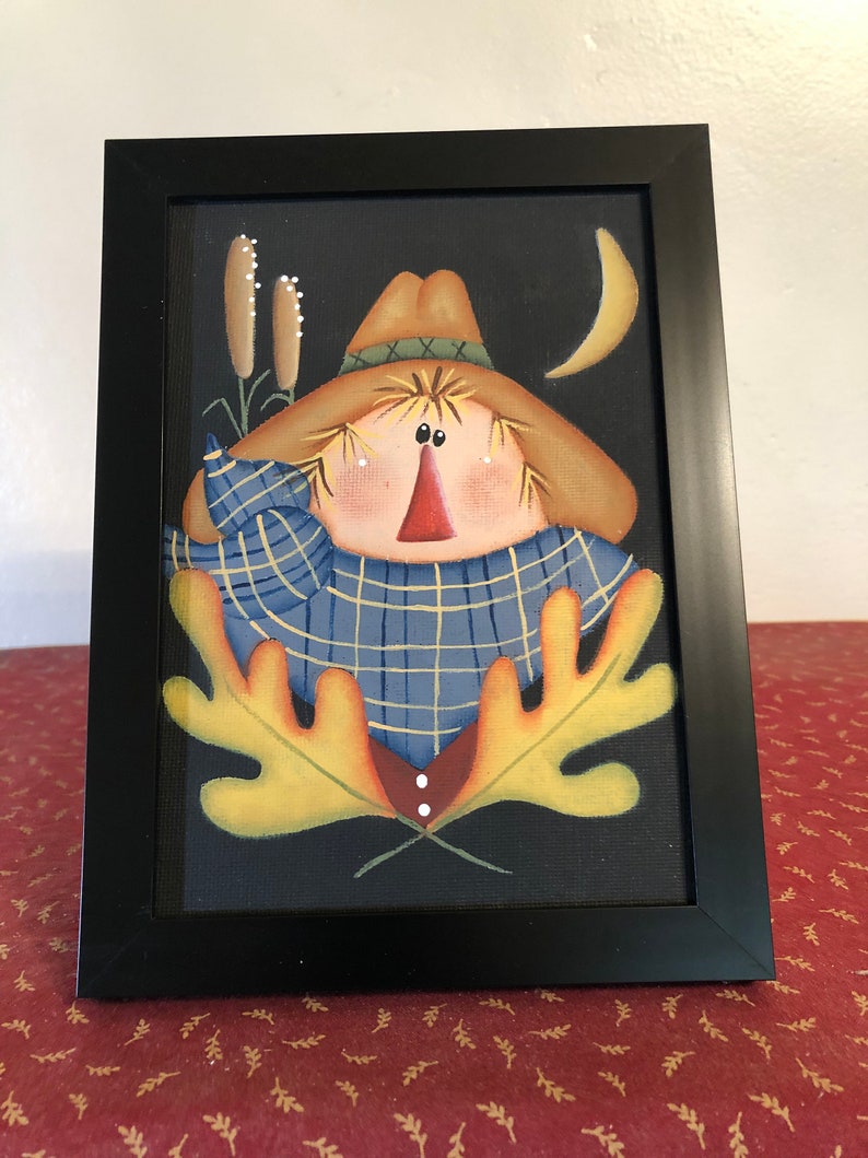 May include: Framed folk art painting of a scarecrow with a straw hat and blue plaid shirt. The scarecrow has a red nose and is surrounded by yellow leaves, cattails, and a crescent moon against a black background. The artwork is in a black frame.