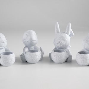 May include: Four small, light gray figurines of cartoon animals, each holding a small, round pot. The figurines include a duck, a turtle, a rabbit, and a cat. The objects are made of a textured material and are arranged in a row.