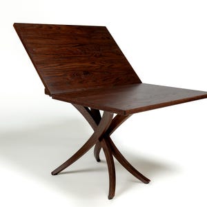 May include: A dark brown wooden table with a unique X-shaped base. The table top is rectangular and has a leaf that can be raised to extend the table's length.